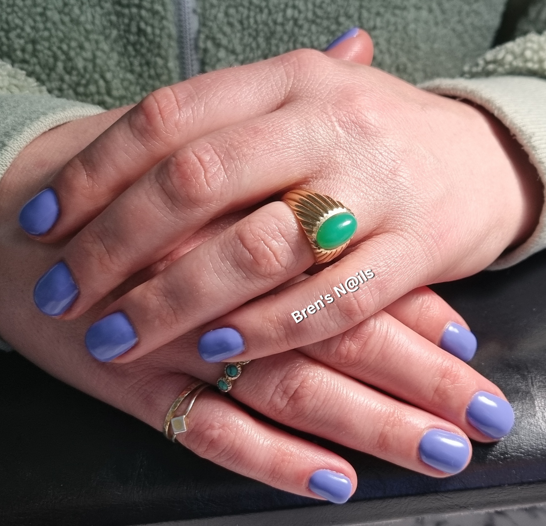 Lavendelblauwe gellak op korte nagels, handen met ringen op een grijze fleece